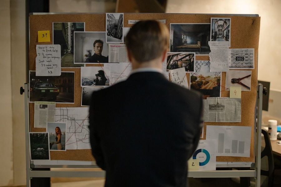 An investigator studying a case board of photographs, maps and handwritten notes.