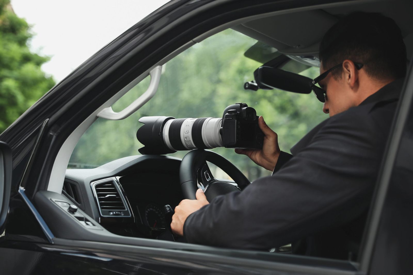 A private investigator seated in a parked vehicle, photographing with a long telephoto lens.